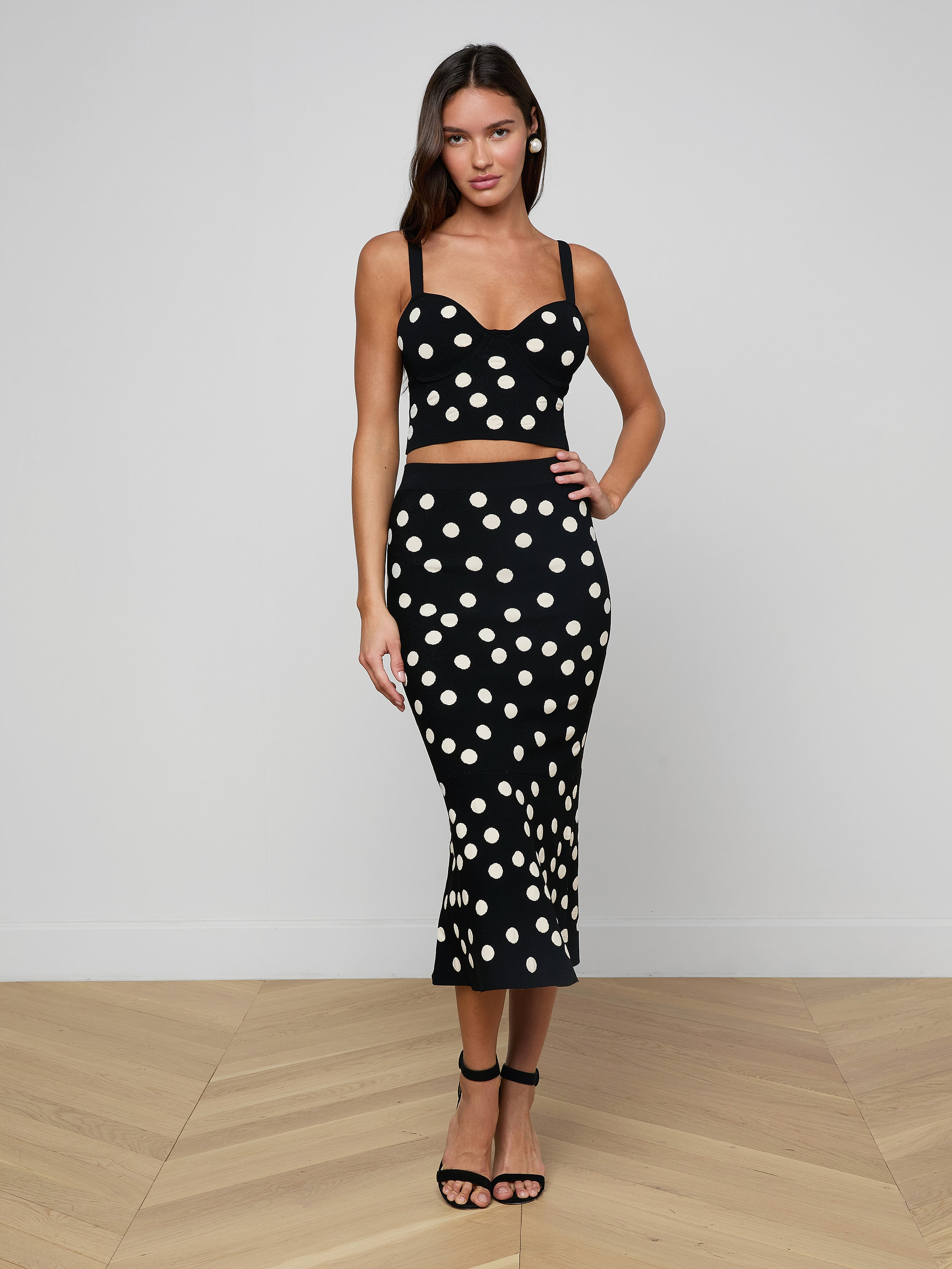 A woman stands against a plain, light background wearing a black, sleeveless crop top paired with the Mae Knit Flare Skirt—a high-waisted, polka dot fit and flare skirt—and black sandals.