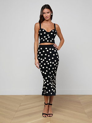 A woman with long dark hair stands indoors against a plain light wall, wearing the Mae Knit Flare Skirt as part of a matching black jacquard polka dot set, paired with black heeled sandals and resting her hand on her hip.