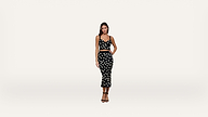 A woman stands against a plain white background wearing a black crop top and matching high-waisted skirt, both with large white polka dots. She has one hand on her hip and is wearing black heeled sandals.