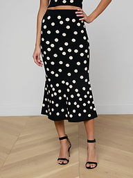A woman wearing the Mae Knit Flare Skirt—featuring bold white polka dots on black—paired with a black sleeveless top and black high-heeled sandals, stands against a plain light background.