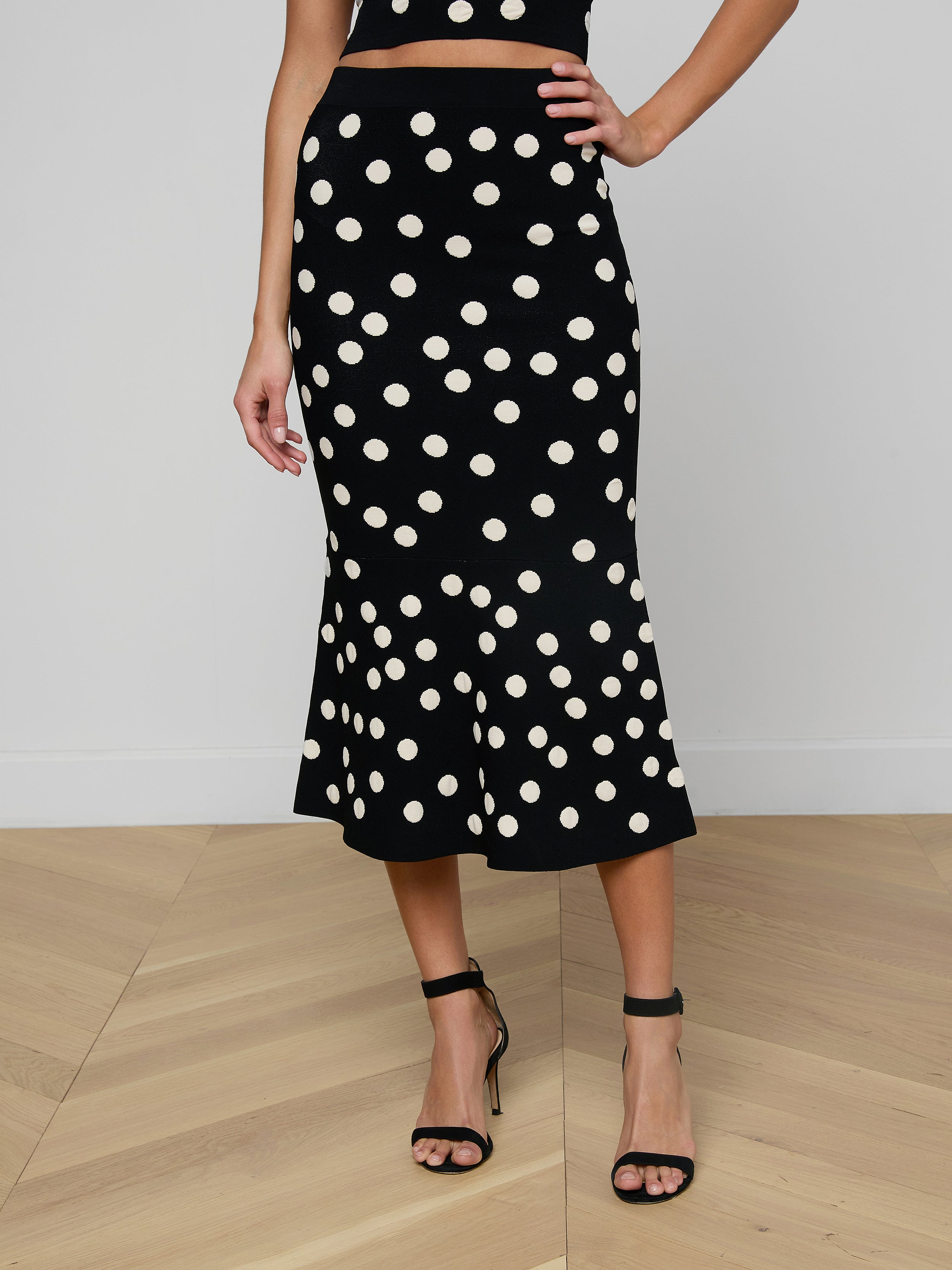 A woman wearing the Mae Knit Flare Skirt—featuring bold white polka dots on black—paired with a black sleeveless top and black high-heeled sandals, stands against a plain light background.