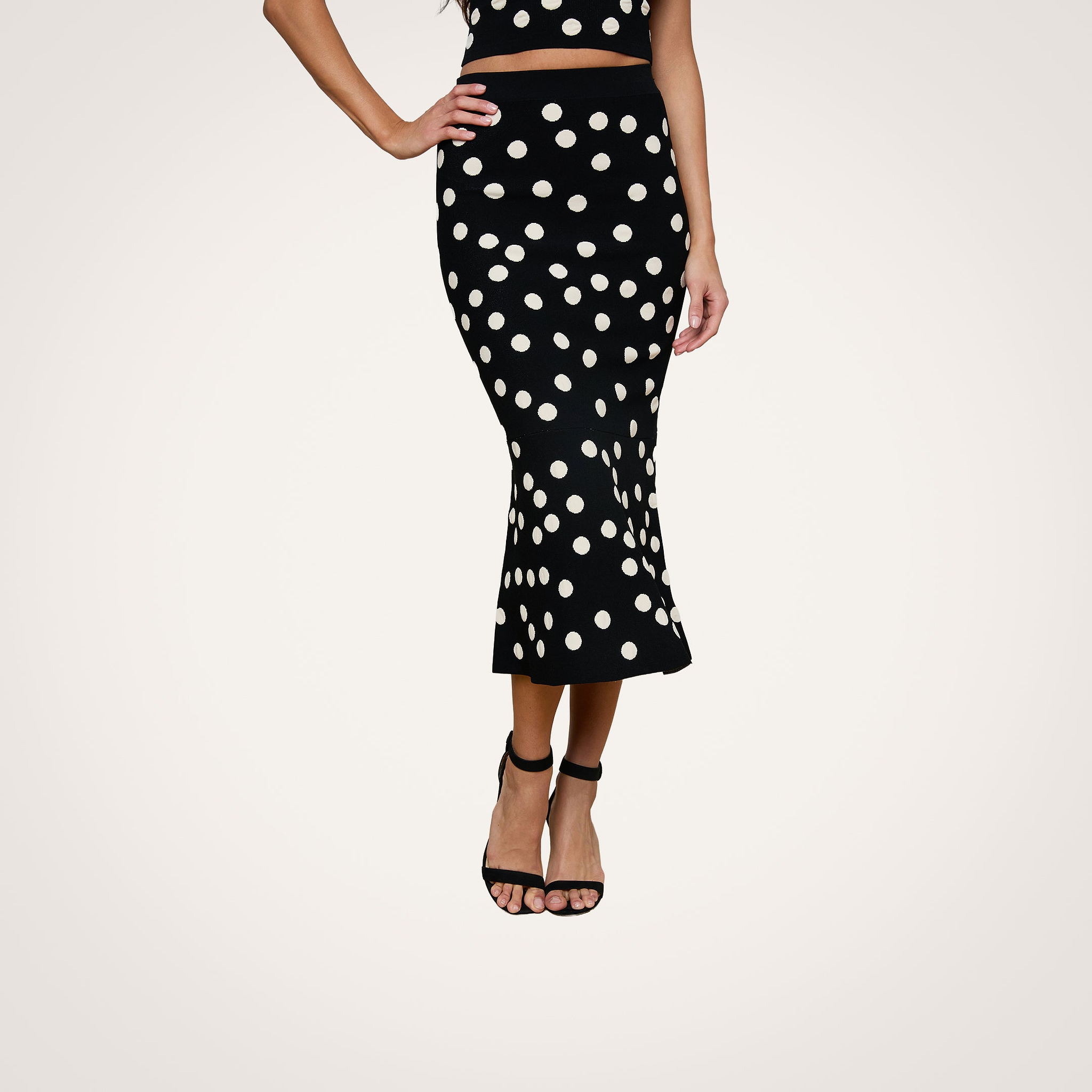 A person wearing the Mae Knit Flare Skirt, a black, high-waisted, ankle-length skirt with white polka dots, paired with a matching crop top and black open-toe high heel sandals. The outfit is photographed against a white background.