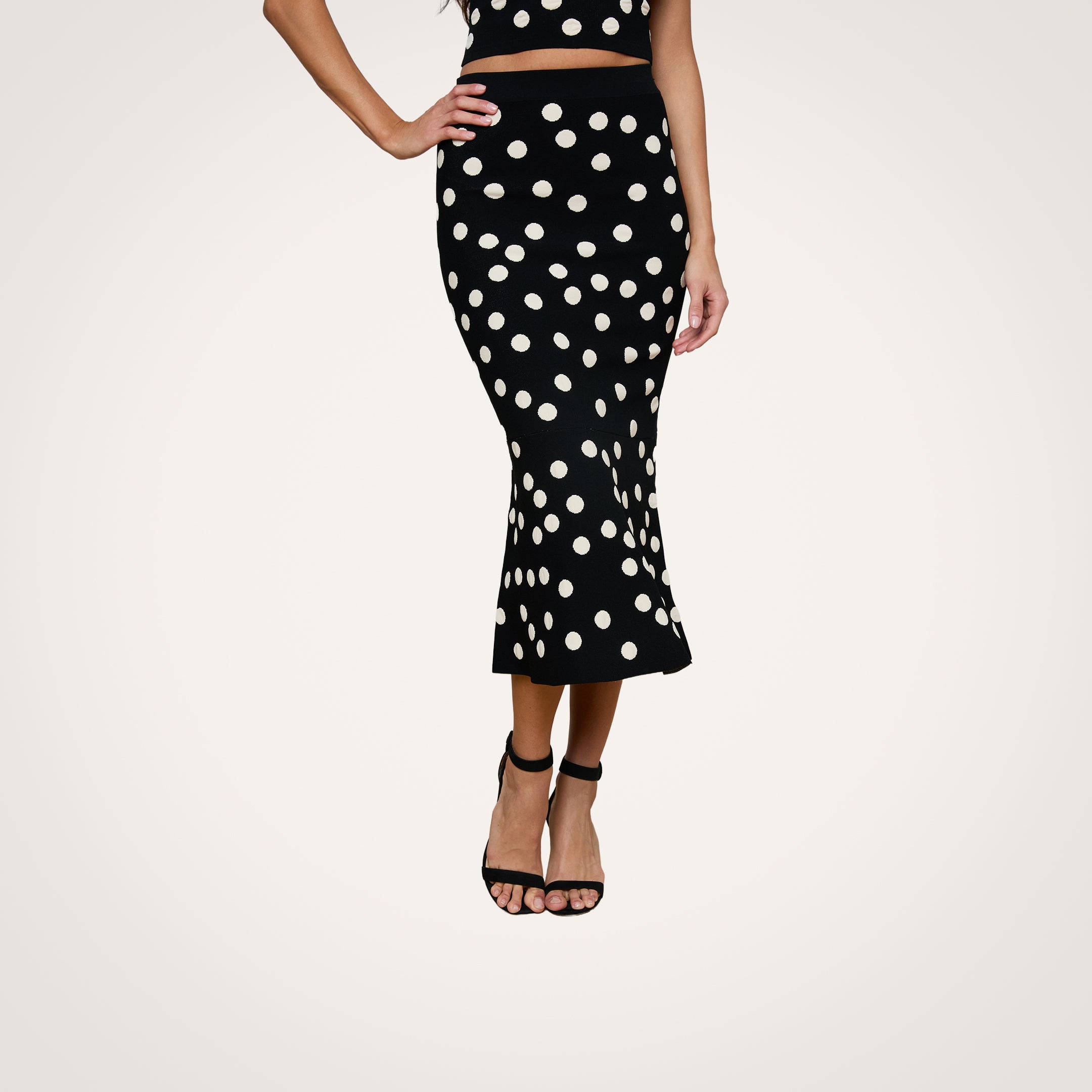 A person wearing the Mae Knit Flare Skirt, a black, high-waisted, ankle-length skirt with white polka dots, paired with a matching crop top and black open-toe high heel sandals. The outfit is photographed against a white background.