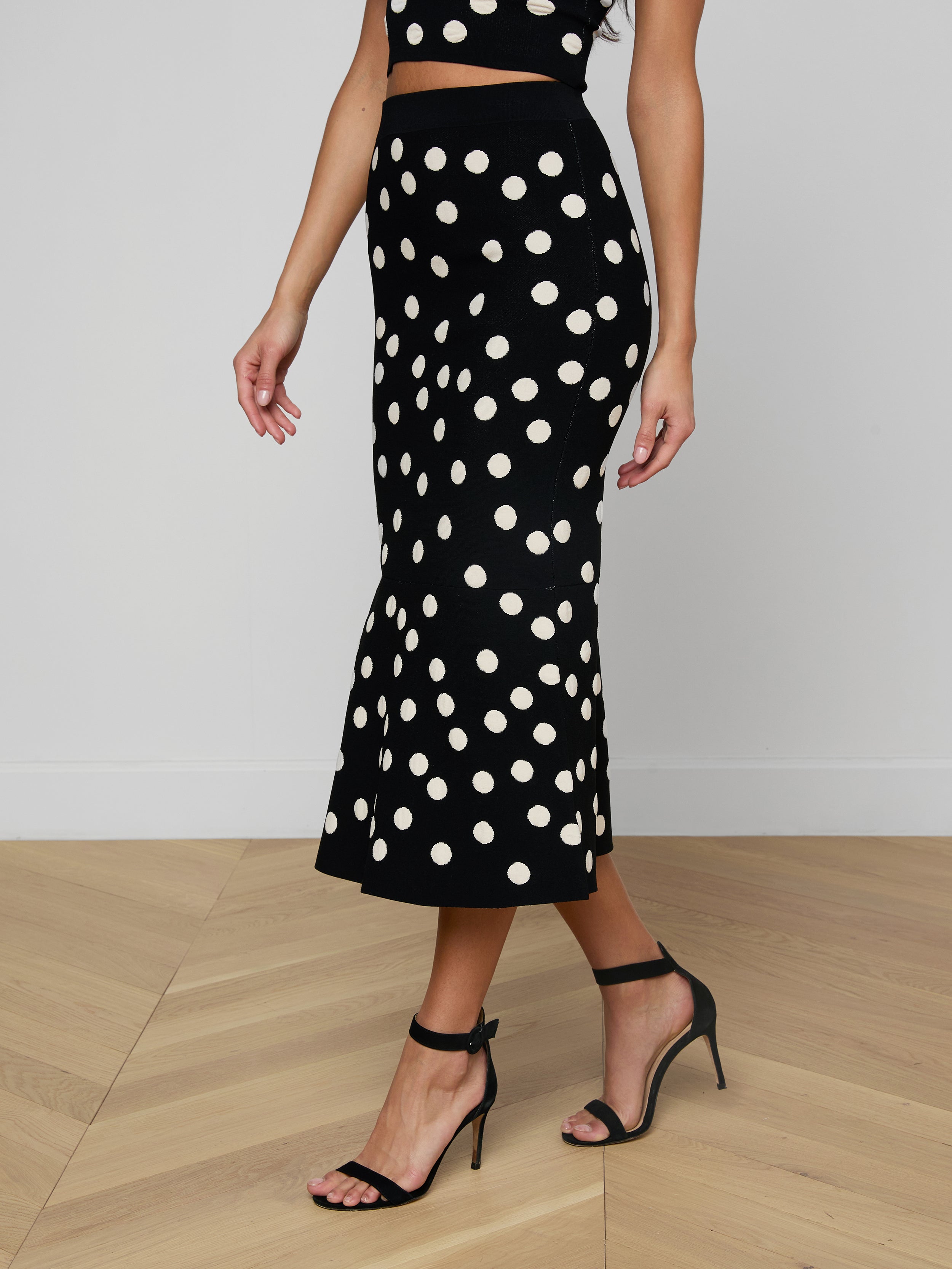 A woman wears the Mae Knit Flare Skirt in black polka dot, paired with coordinating high-heeled sandals and a matching polka dot top. The fit and flare design of the skirt creates a timeless silhouette against the plain white background.