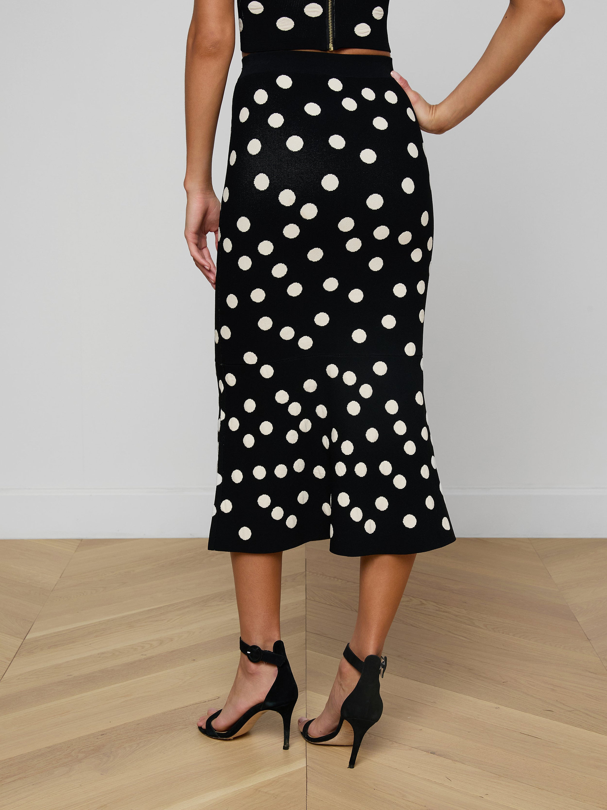 A woman stands with one hand on her hip, wearing the Mae Knit Flare Skirt in black with white polka dots and black high-heeled sandals. The photo is taken from behind, showing only her lower back and legs.