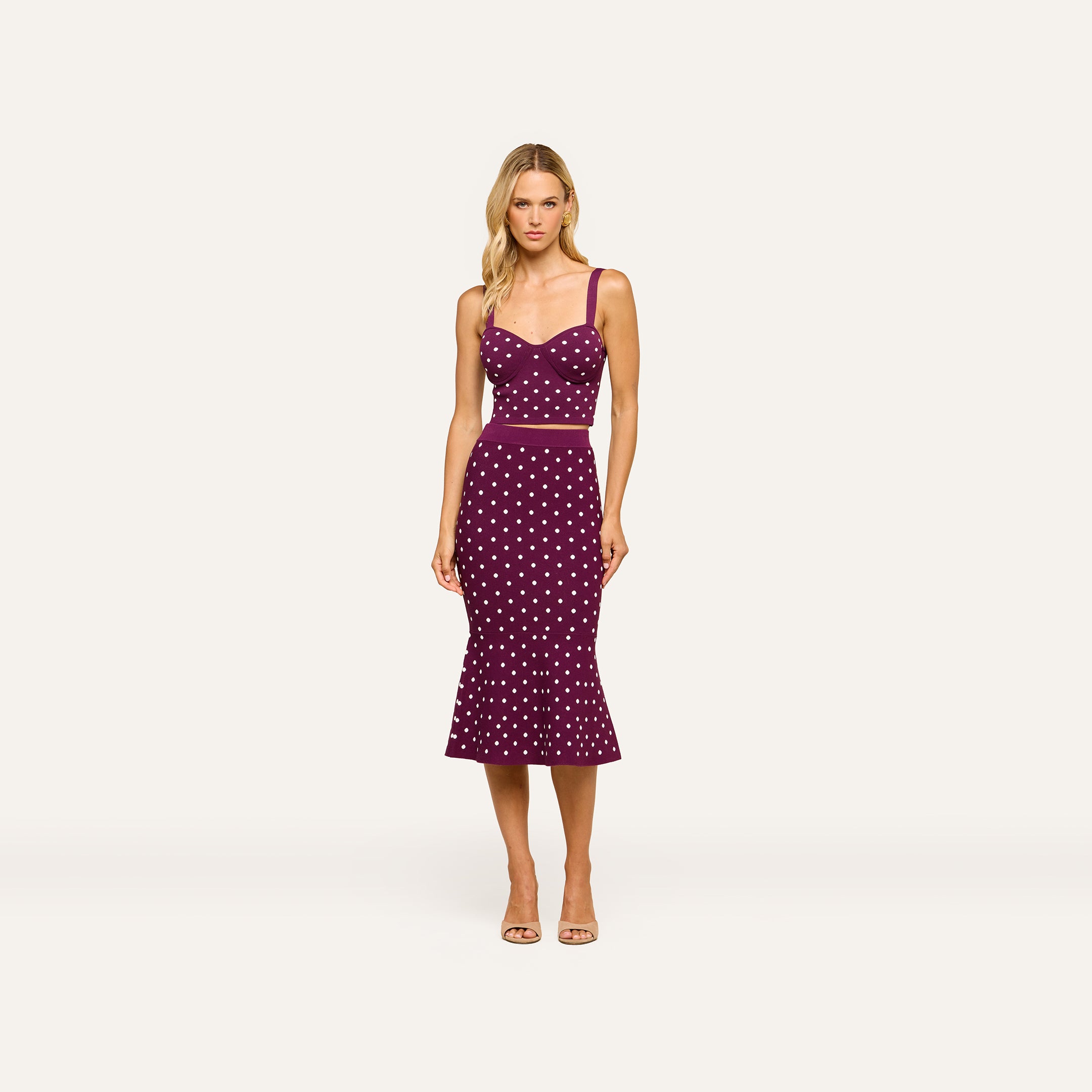 A woman stands facing forward against a plain background, wearing the Mae Knit Flare Skirt — a sleeveless, fitted purple dress with white polka dots and a flared hem that creates a flattering silhouette. She has blonde hair and is paired with open-toed heels.