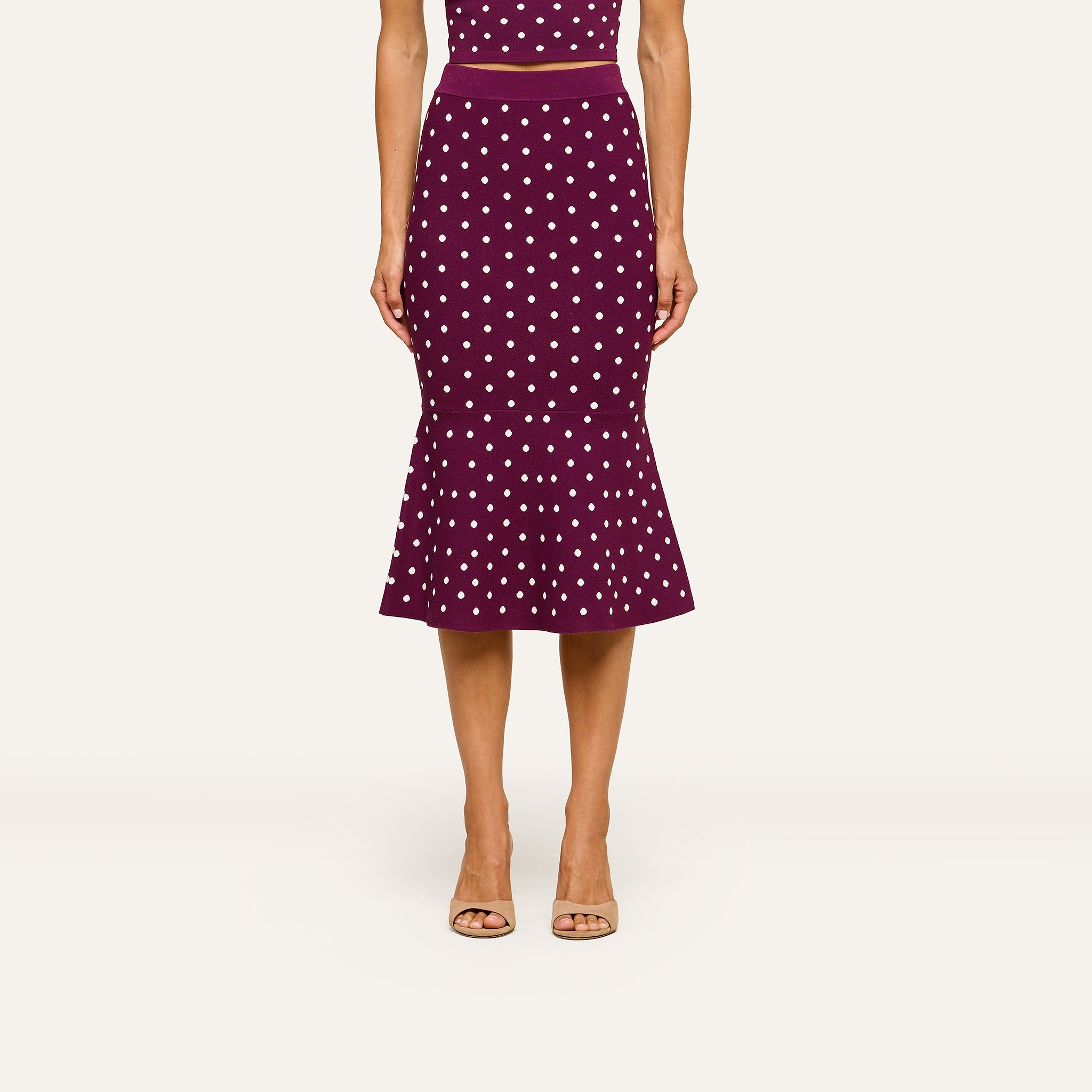 A woman wearing a sleeveless, fitted purple dress paired with the Mae Knit Flare Skirt and beige open-toe heels stands against a plain light background.