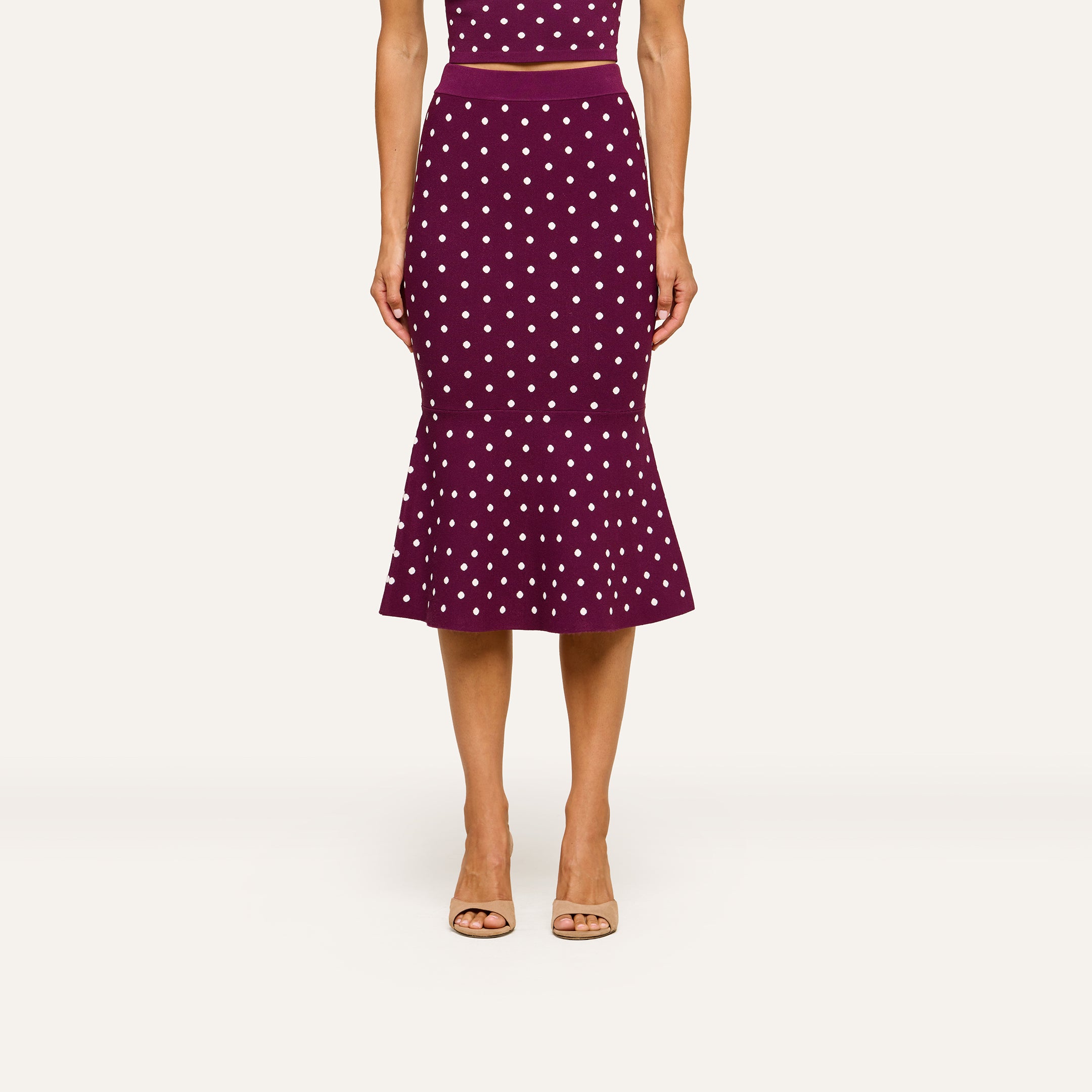 A woman wearing a sleeveless, fitted purple dress paired with the Mae Knit Flare Skirt and beige open-toe heels stands against a plain light background.