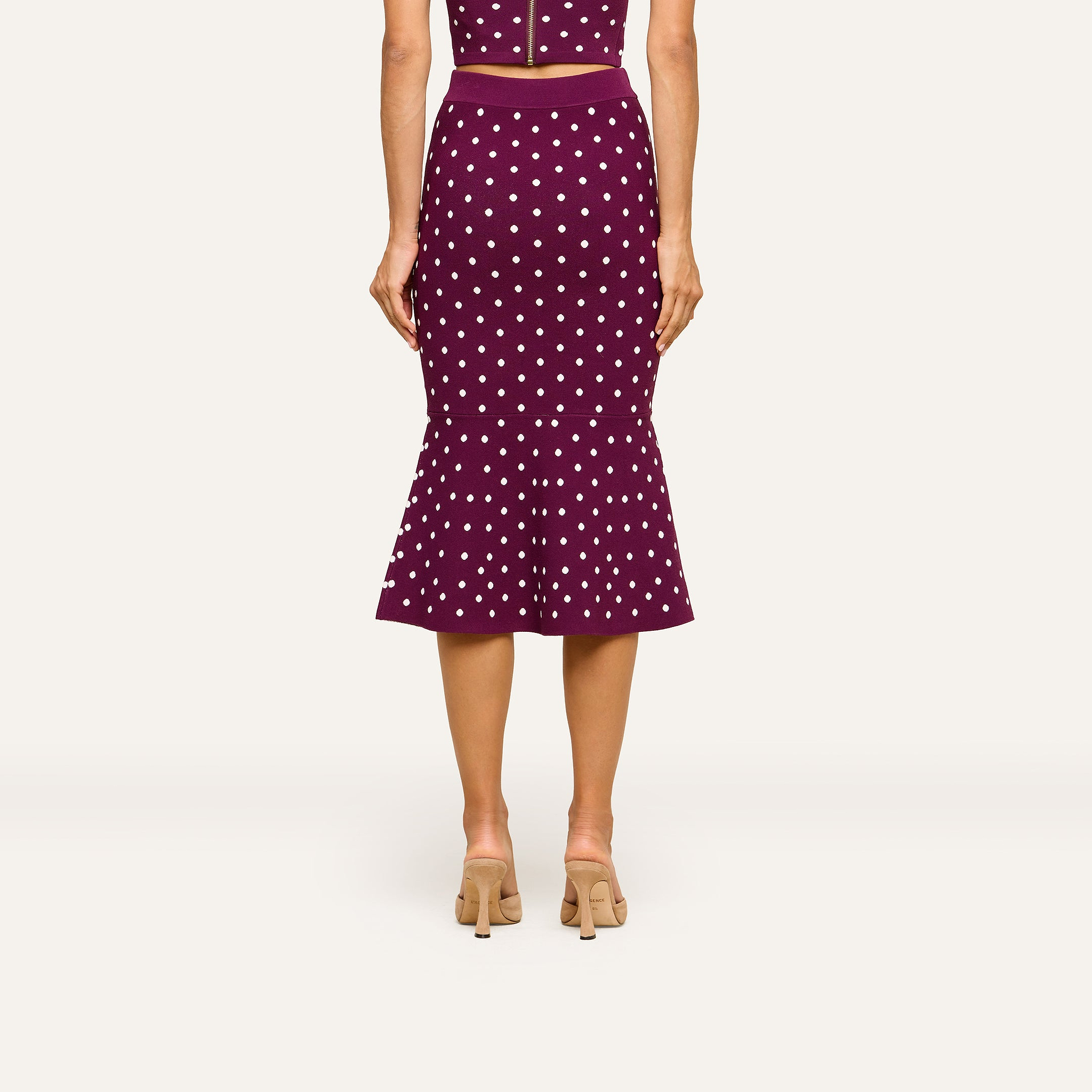 A woman stands facing away, showcasing the Mae Knit Flare Skirt—a purple polka dot midi with a flared hem—paired with tan high-heeled shoes. Only her lower back, skirt, and legs are visible against a plain background.