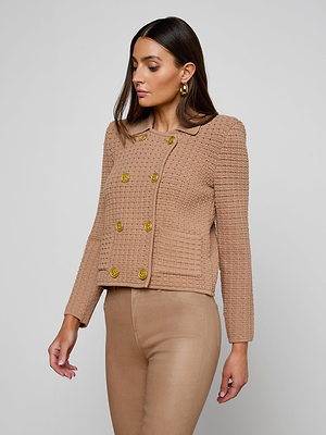 A woman with long brown hair models the Charlie Knit Jacket—a textured, camel-colored style with gold buttons—paired with tan pants against a plain light gray background. She looks slightly down and to the side.