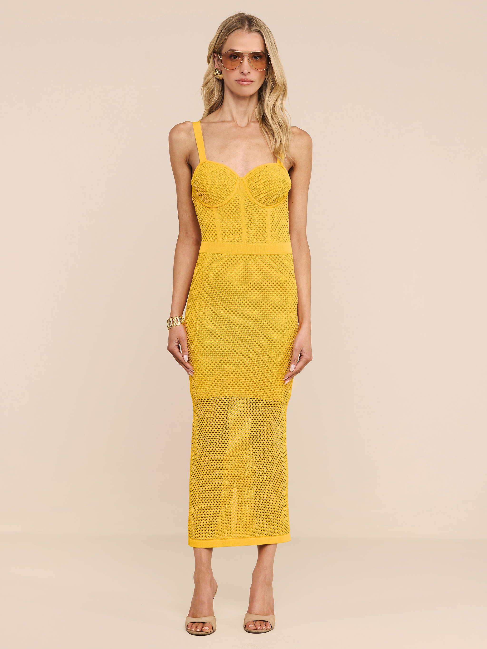 Una donna dai capelli biondi e mossi indossa il Michelle Knit Dress, un abito aderente in maglia di colore giallo brillante con gonna in rete e top in stile bustier, abbinato a occhiali da sole, un braccialetto e sandali con tacco beige, su uno sfondo semplice.