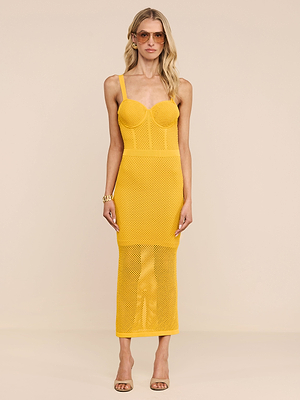 A woman with wavy blonde hair wears the Michelle Knit Dress, a bright yellow fitted knit dress with a mesh skirt and bustier-style top, paired with sunglasses, a bracelet, and beige heeled sandals against a plain background.