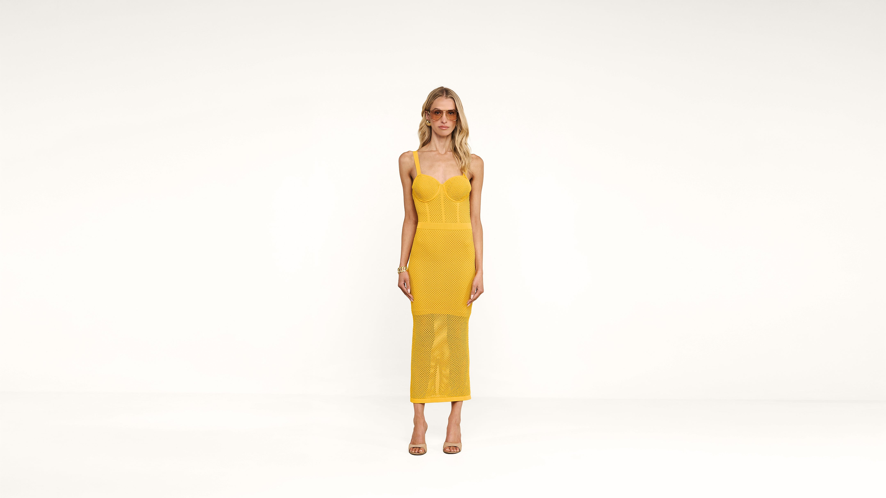 A woman stands against a plain white background, wearing a yellow fitted, sleeveless midi dress with spaghetti straps and open-toed heels.