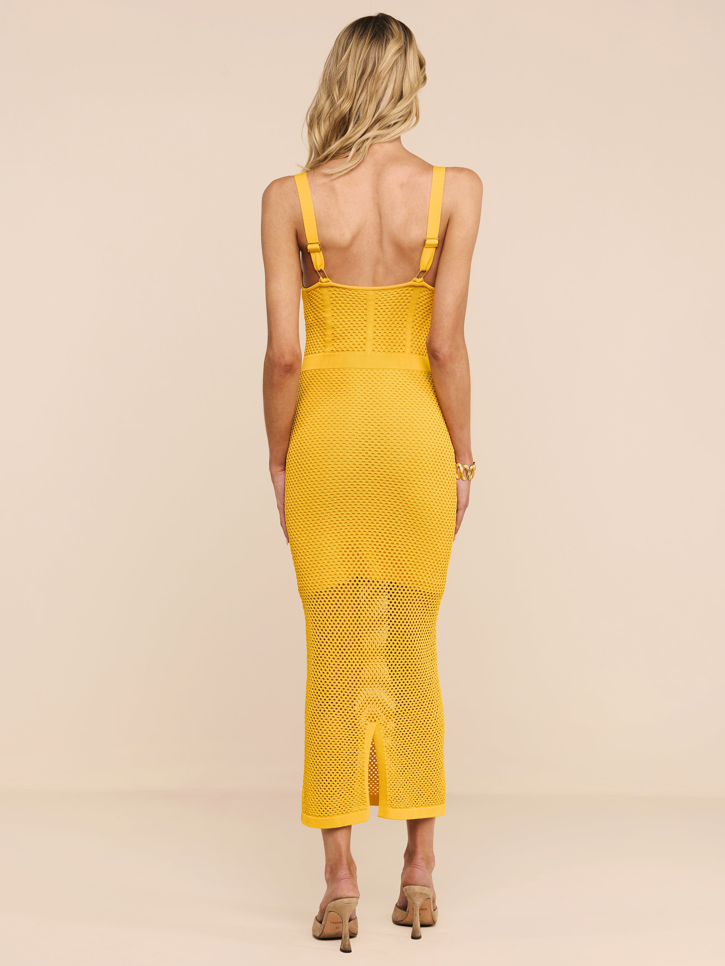 Una donna dai capelli biondi indossa labito in maglia Michelle di colore giallo e sandali con tacco beige, in piedi di spalle alla fotocamera su uno sfondo beige a tinta unita.