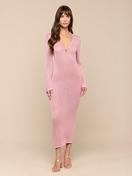 A woman wearing the Sydney Knit Dress—a long-sleeve, light pink, form-fitting maxi—stands against a plain white background, looking forward. She pairs the dress with clear heeled sandals and has long brown hair with bangs.