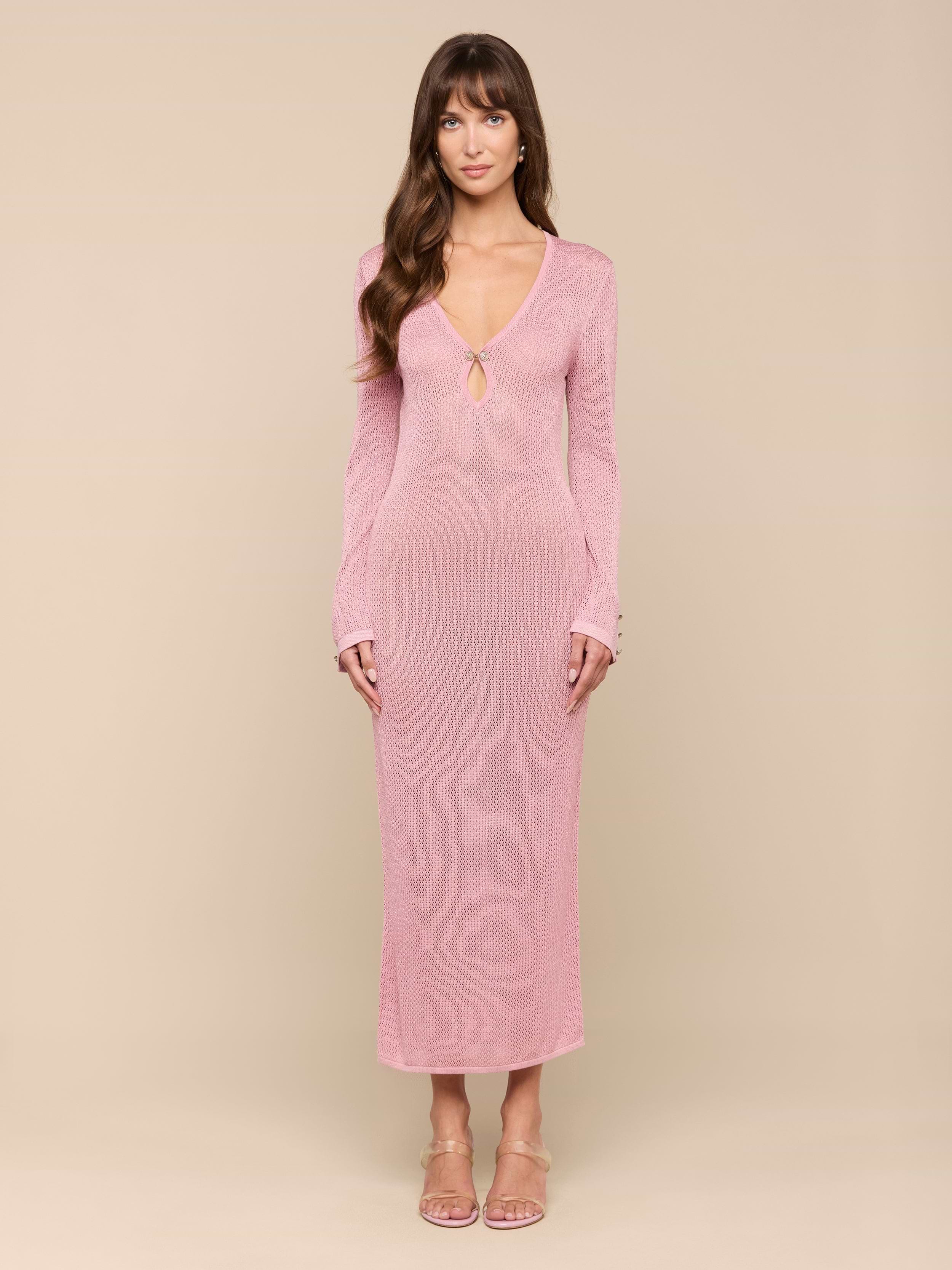 A woman stands facing forward against a plain white background, wearing the Sydney Knit Dress—a long-sleeve, pink maxi dress with a V-neckline and keyhole detail in pointelle knit—paired with open-toe heels.