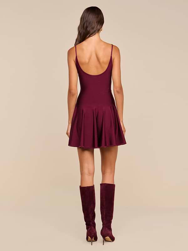A woman with long brown hair, wearing the Lana Flare Knit Dress—a sleeveless burgundy stretch knit mini dress with adjustable straps—and matching knee-high boots, stands facing away against a plain white background.