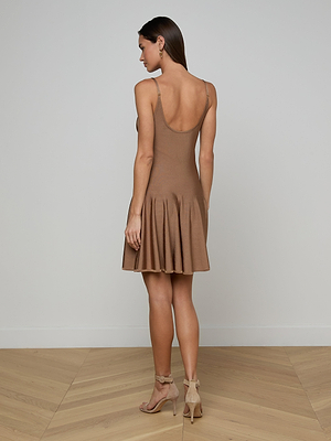 A woman stands indoors on a wooden floor, facing away. She wears the sleeveless, tan Lana Knit Dress with a knee-length flared skirt and beige high-heeled sandals. The background is a plain light-colored wall.