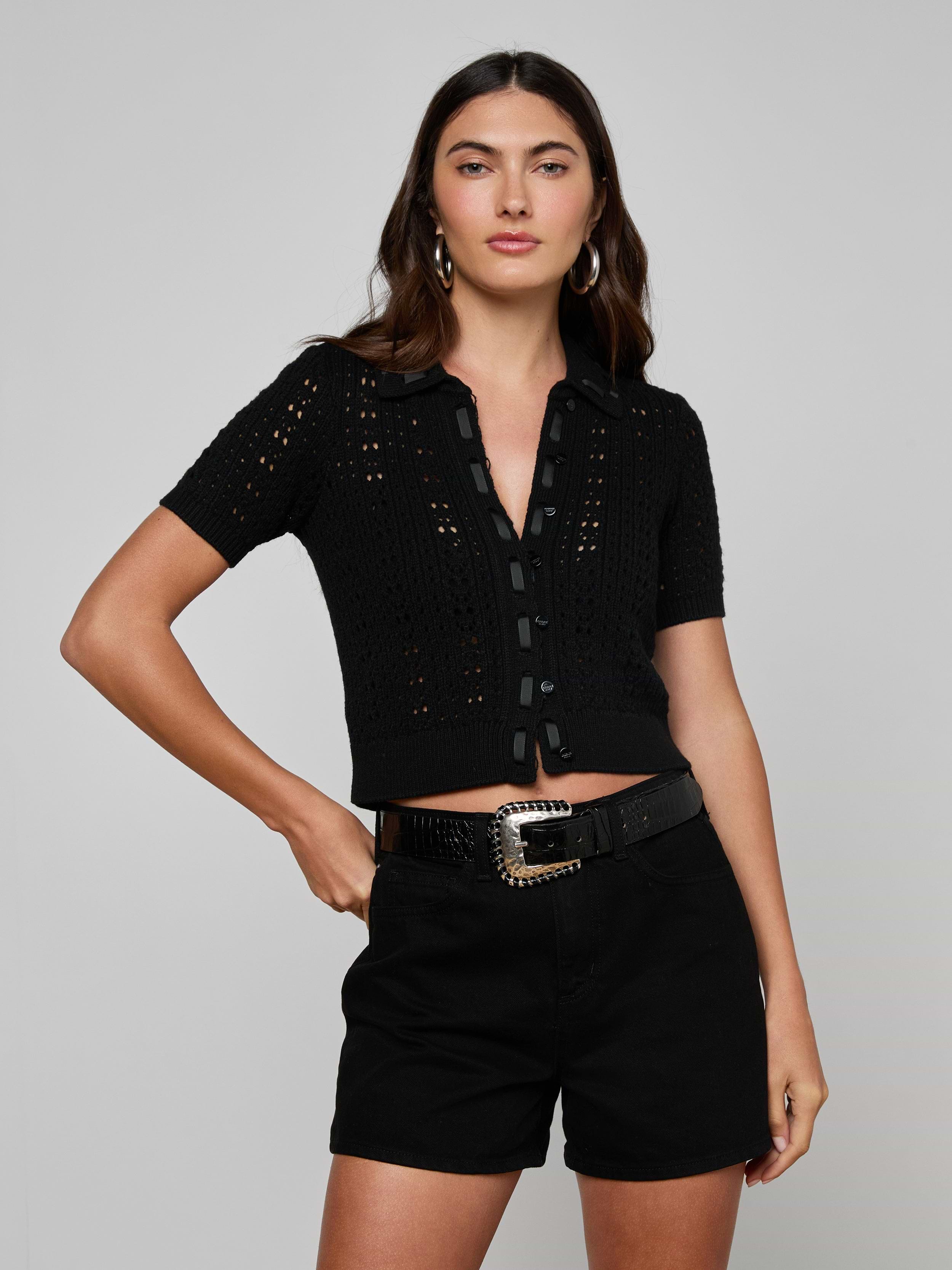 A woman stands against a light background, wearing the Ninette Knit Top—a black short-sleeve pointelle knit top with buttons—paired with black shorts, a black belt featuring a decorative buckle, and hoop earrings. One hand rests on her hip, exuding Parisian flair.