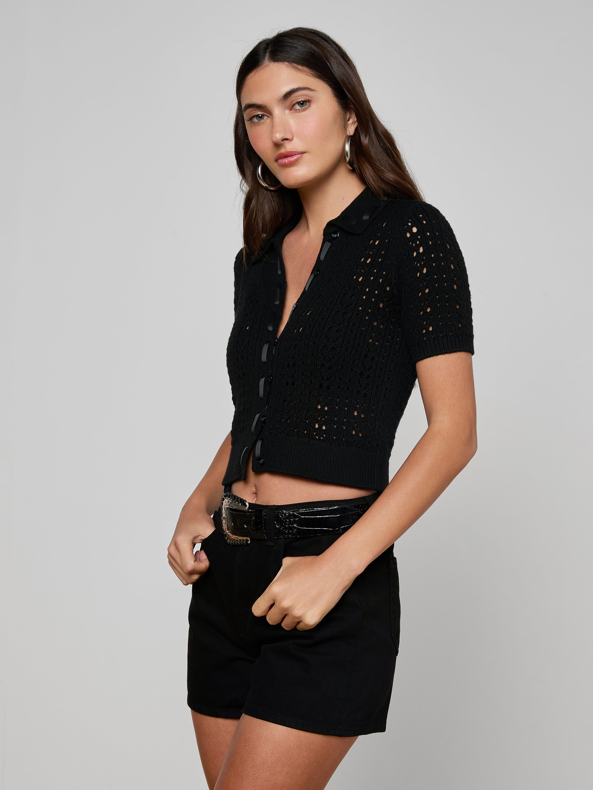 Wearing the Ninette Knit Top—a black short-sleeve, button-up crochet design with grosgrain ribbon trim—a woman poses with black belted shorts against a plain, light background.