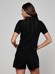 A woman with long brown hair stands facing away, wearing the Ninette Knit Top—a short-sleeve black pointelle knit top—paired with black shorts. Her hands rest on her hips, and she wears large hoop earrings against a plain light background.