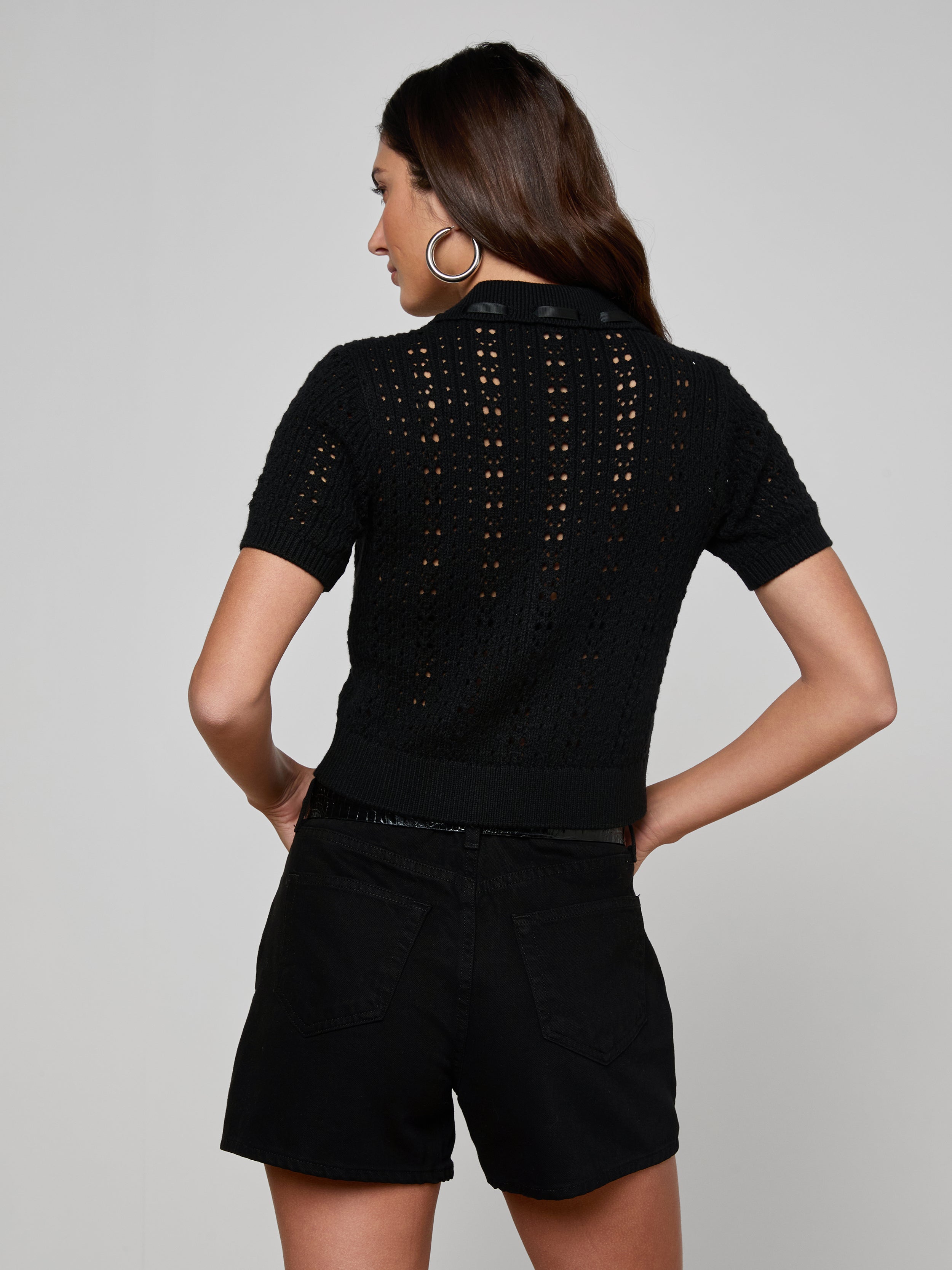 A woman with long brown hair stands facing away, wearing the Ninette Knit Top—a short-sleeve black pointelle knit top—paired with black shorts. Her hands rest on her hips, and she wears large hoop earrings against a plain light background.