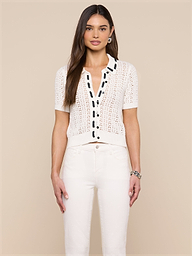 A woman with long brown hair wears the Ninette Knit Top, a short-sleeve white shirt with black button trim, paired with white pants. She stands facing forward against a plain, light background.