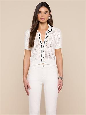 A woman with long brown hair stands against a beige background wearing the Ninette Knit Top— a white short-sleeved pointelle cardigan with black grosgrain ribbon trim—paired with white pants, earrings, and a bracelet for Parisian flair.