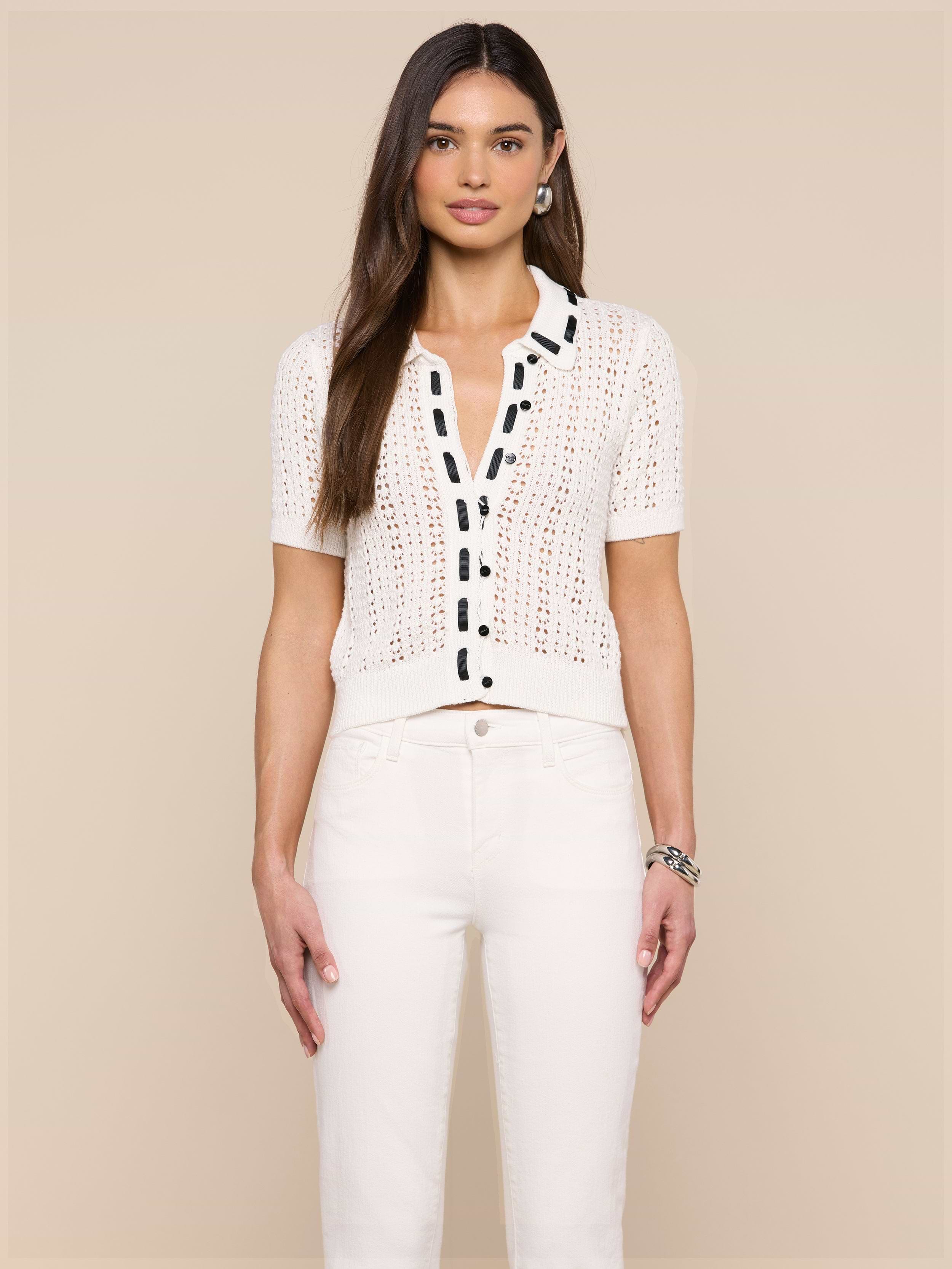 A woman with long brown hair wears the Ninette Knit Top, a short-sleeve white shirt with black button trim, paired with white pants. She stands facing forward against a plain, light background.