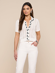 A woman stands against a plain background, wearing the Ninette Knit Top—a white short-sleeve, button-up pointelle knit cardigan with black grosgrain ribbon trim—paired with high-waisted white pants. Her right hand rests in her pocket, and she has long brown hair.