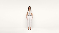 A woman with long brown hair stands against a plain white background, wearing a white short-sleeve blouse, white pants, a black belt, and black high-heeled sandals.