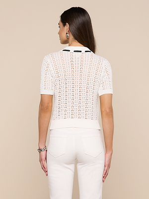 A woman with brown hair stands facing away, wearing the Ninette Knit Top—a white pointelle cardigan with a black collar—paired with white high-waisted pants, hoop earrings, and bracelets against a beige background for Parisian-inspired style.