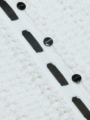 Close-up of the Ninette Knit Top, a textured white pointelle cardigan with vertical grosgrain ribbon trim and several round black buttons, each engraved for an elegant Parisian-inspired finish.