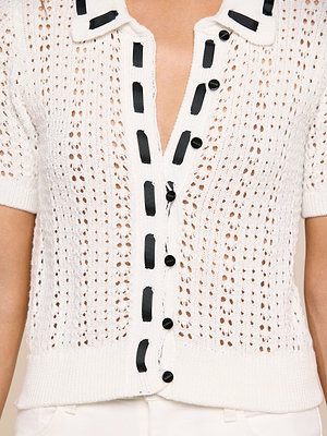 Someone wearing the Ninette Knit Top—a white, short-sleeve pointelle knit cardigan with a deep V-neck, black buttons, grosgrain ribbon trim on the placket and collar, and an open airy pattern that exudes Parisian flair.