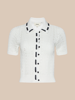 The Ninette Knit Top is a white short-sleeve pointelle cardigan with black buttons and trim along the placket and collar, shown on a beige background for a touch of Parisian flair.