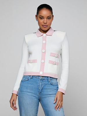 A woman in the Nomi Satin Back Cardigan, a white piece with pink contrast trim and buttons, pairs it with blue jeans and stands confidently before a plain light gray background.