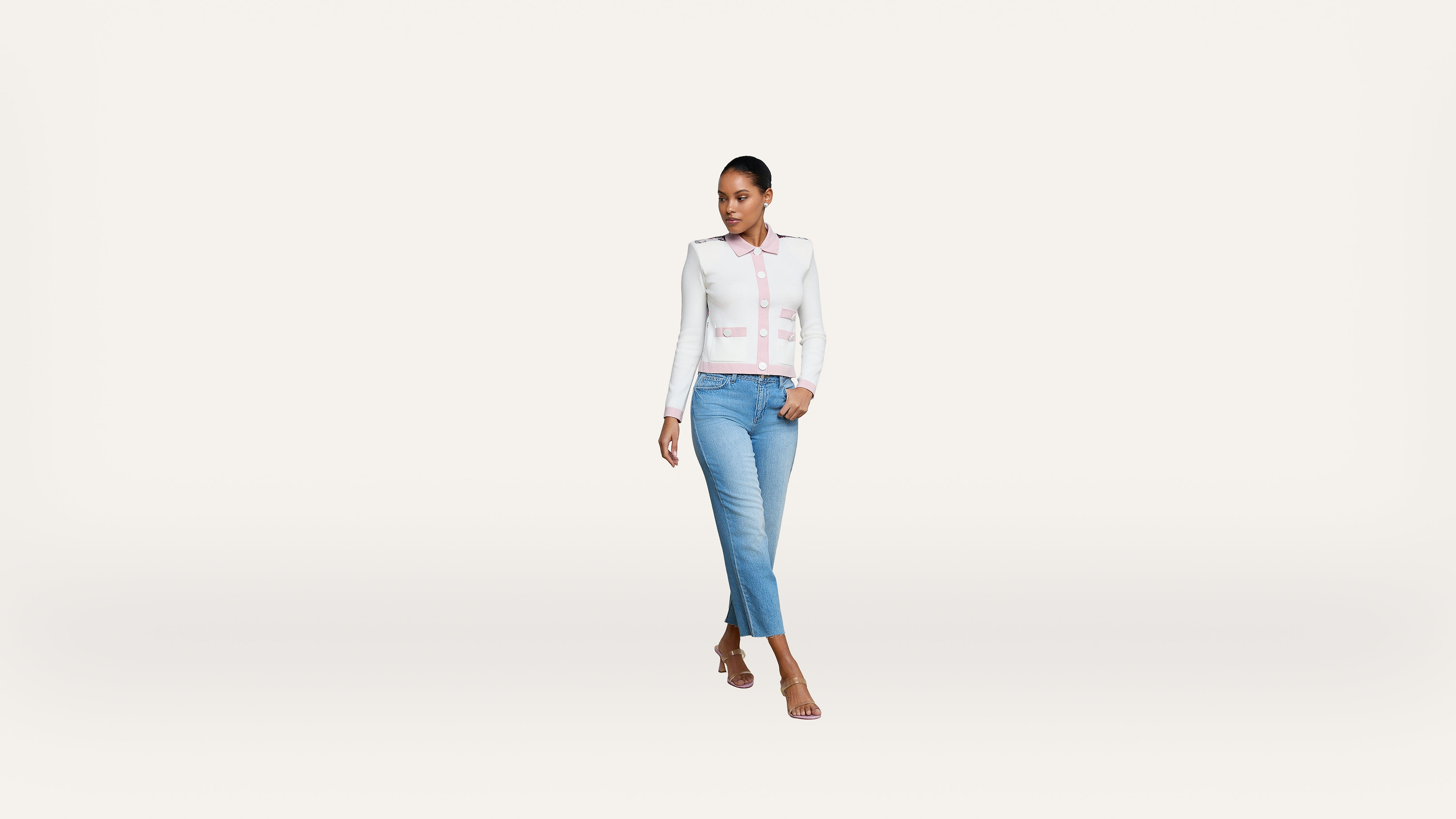A woman wearing a white jacket, light pink shirt, blue jeans, and heels walks confidently against a plain white background.