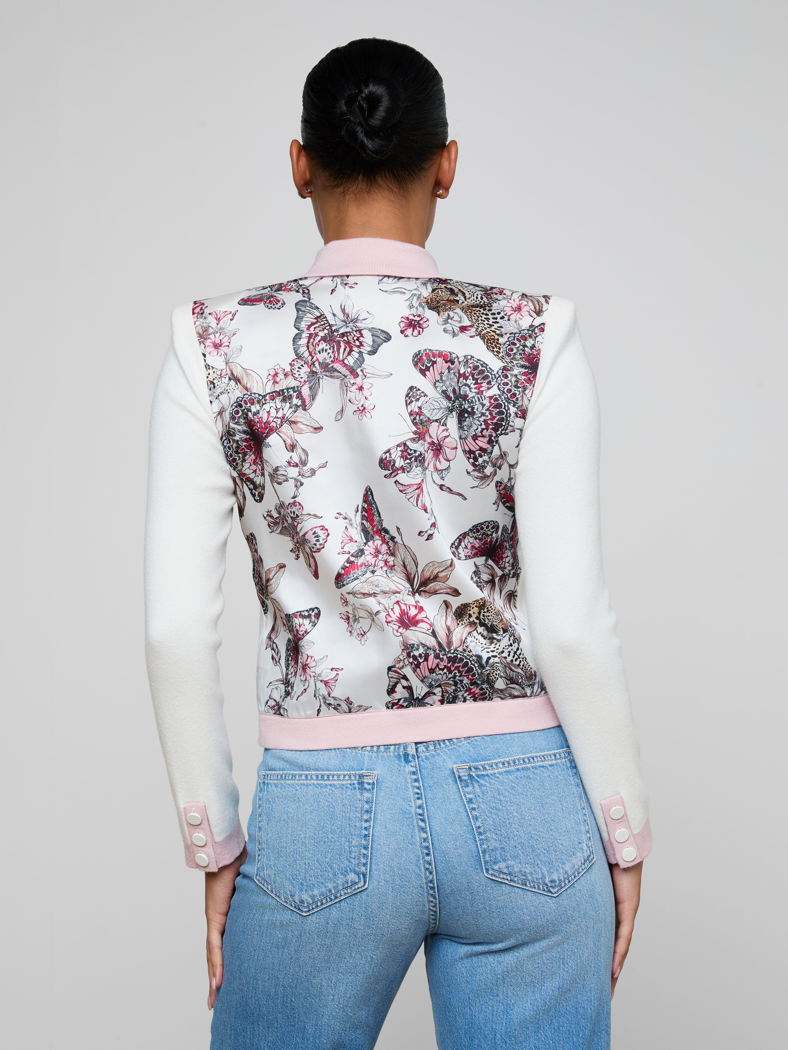 A woman with her hair in a bun is shown from the back against a plain light background, wearing the Nomi Satin Back Cardigan featuring statement buttons and white sleeves, paired with light blue jeans.