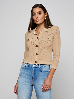 A woman with long brown hair models the Risa Collared Cardigan, a neutral tan pointelle knit with front pockets, paired with blue jeans. She poses against a plain light gray background.
