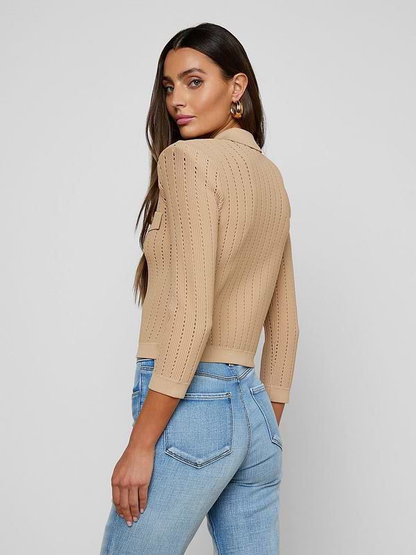 A woman stands sideways and looks over her shoulder, wearing the Risa Collared Cardigan—a beige pointelle knit sweater with three-quarter sleeves—paired with light blue jeans, against a plain light background.