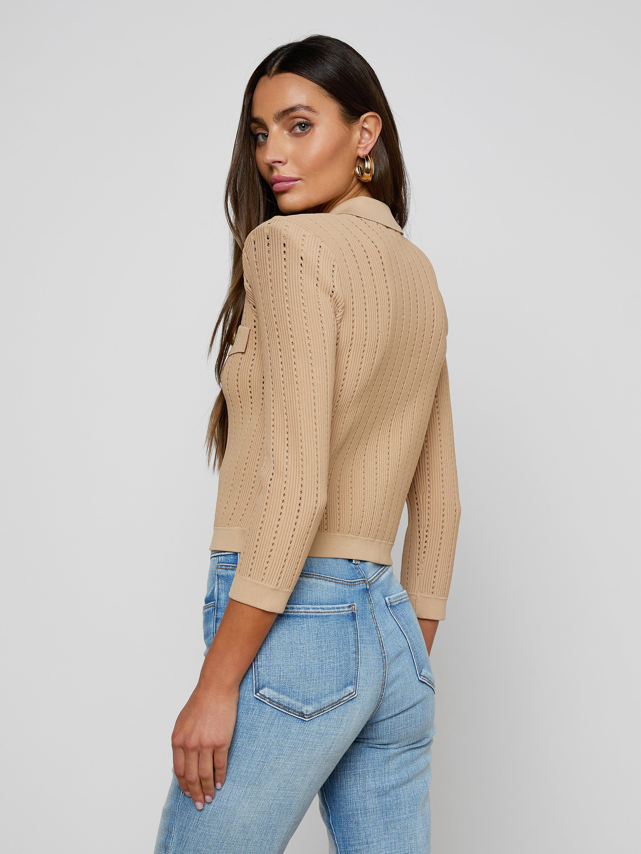 A woman stands sideways and looks over her shoulder, wearing the Risa Collared Cardigan—a beige pointelle knit sweater with three-quarter sleeves—paired with light blue jeans, against a plain light background.