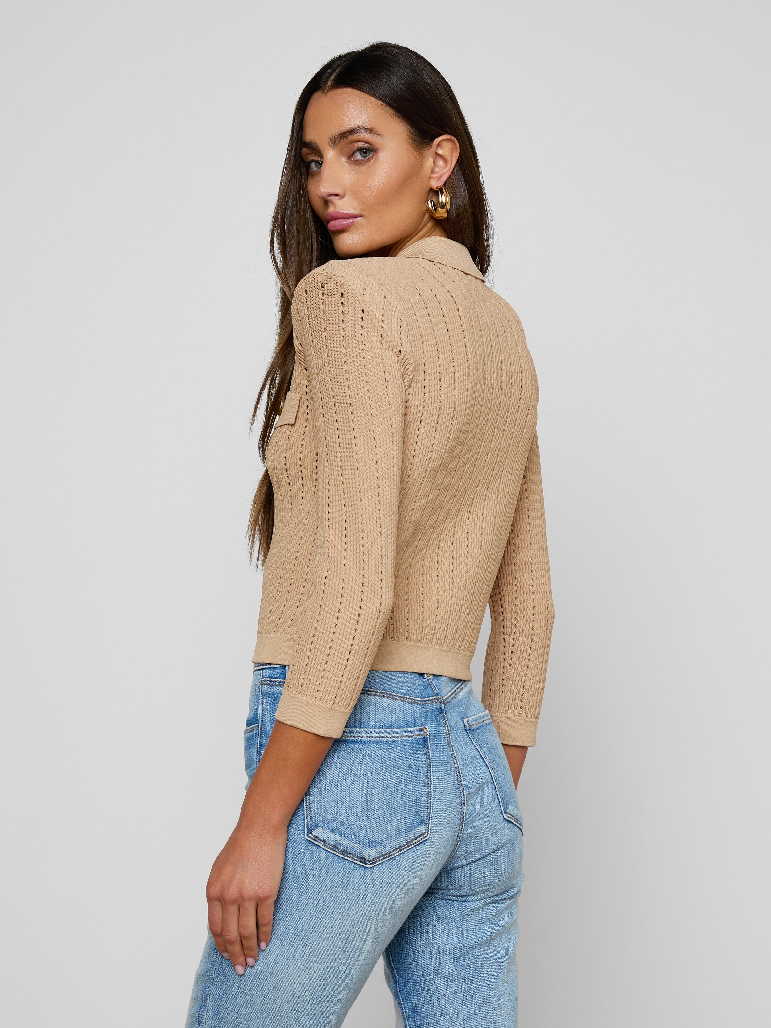 A woman stands sideways and looks over her shoulder, wearing the Risa Collared Cardigan—a beige pointelle knit sweater with three-quarter sleeves—paired with light blue jeans, against a plain light background.