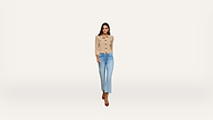 Woman wearing a beige button-up cardigan, light blue jeans, and brown heels walks forward against a plain white background.