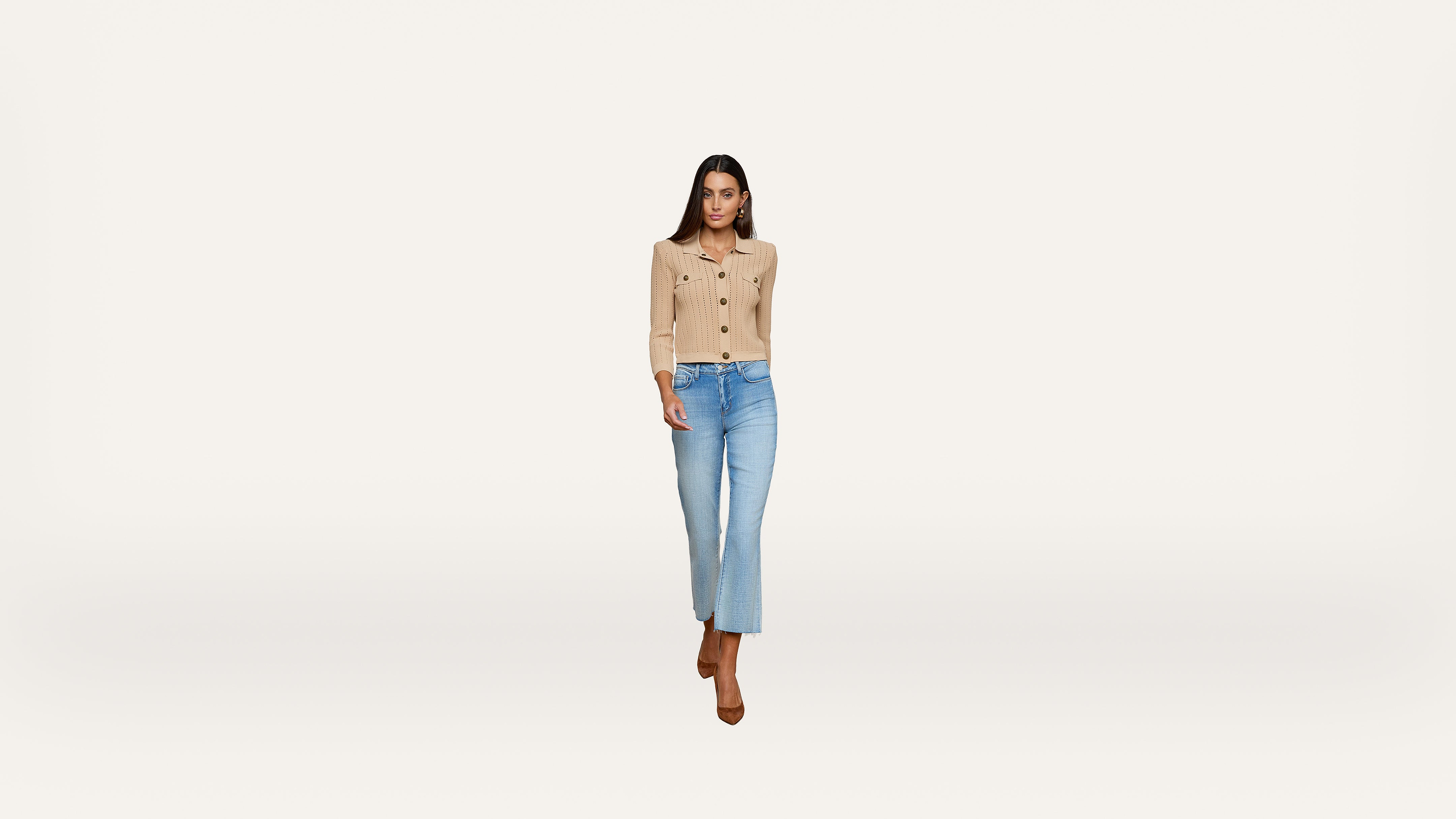 Woman wearing a beige button-up cardigan, light blue jeans, and brown heels walks forward against a plain white background.