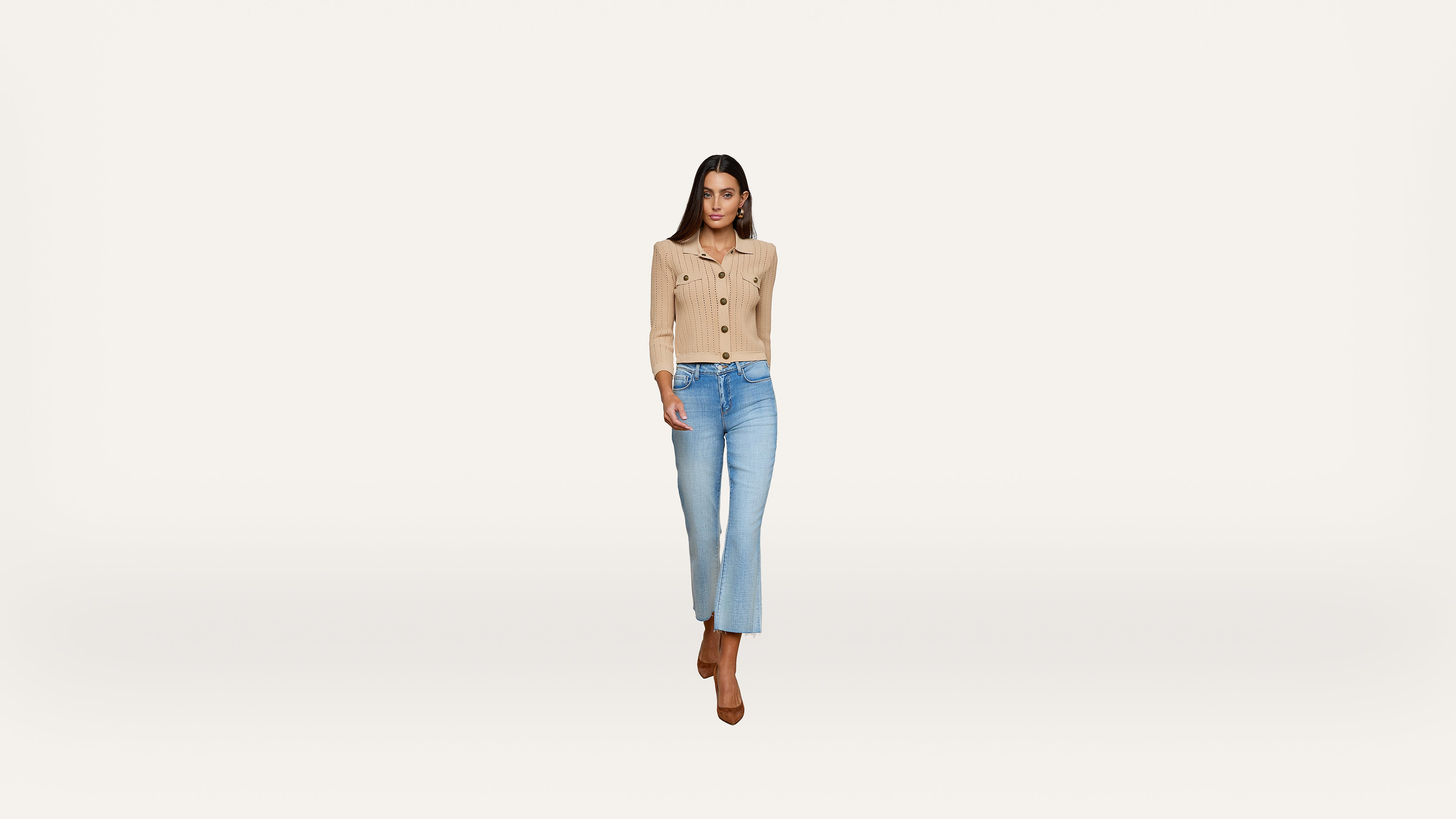 Woman wearing a beige button-up cardigan, light blue jeans, and brown heels walks forward against a plain white background.