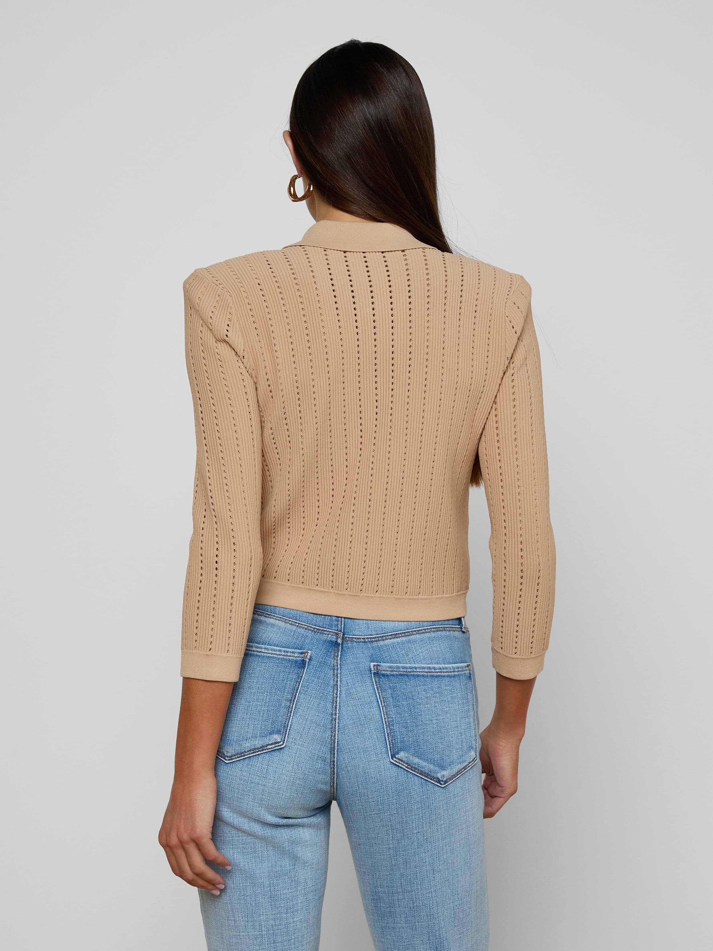 A woman with long brown hair, wearing the Risa Collared Cardigan in beige and light blue jeans, stands facing away from the camera against a plain light background.
