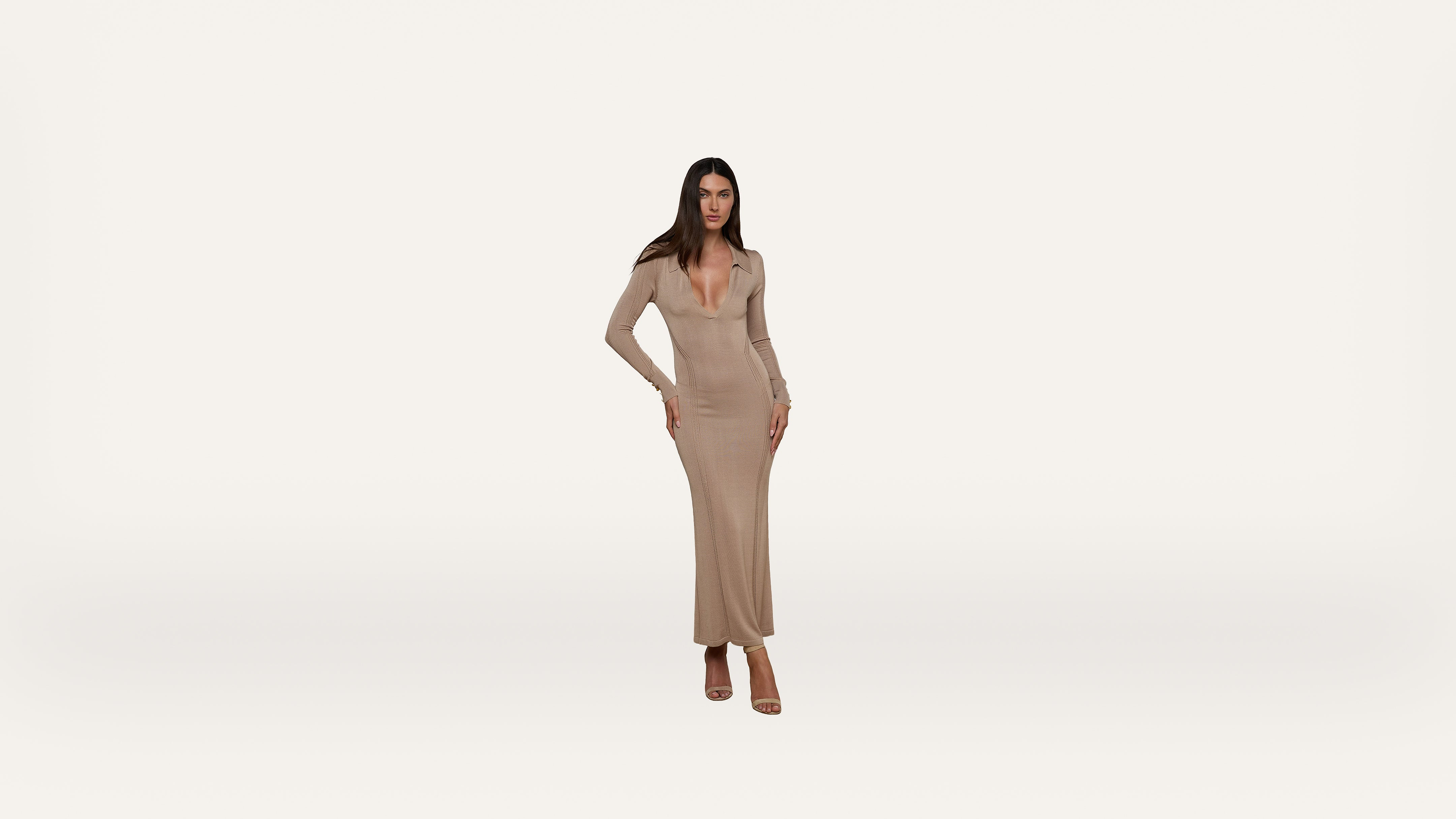 A woman stands confidently in a long, fitted, beige dress with long sleeves and a deep neckline against a plain white background. She is wearing open-toed heels and has long, straight hair.