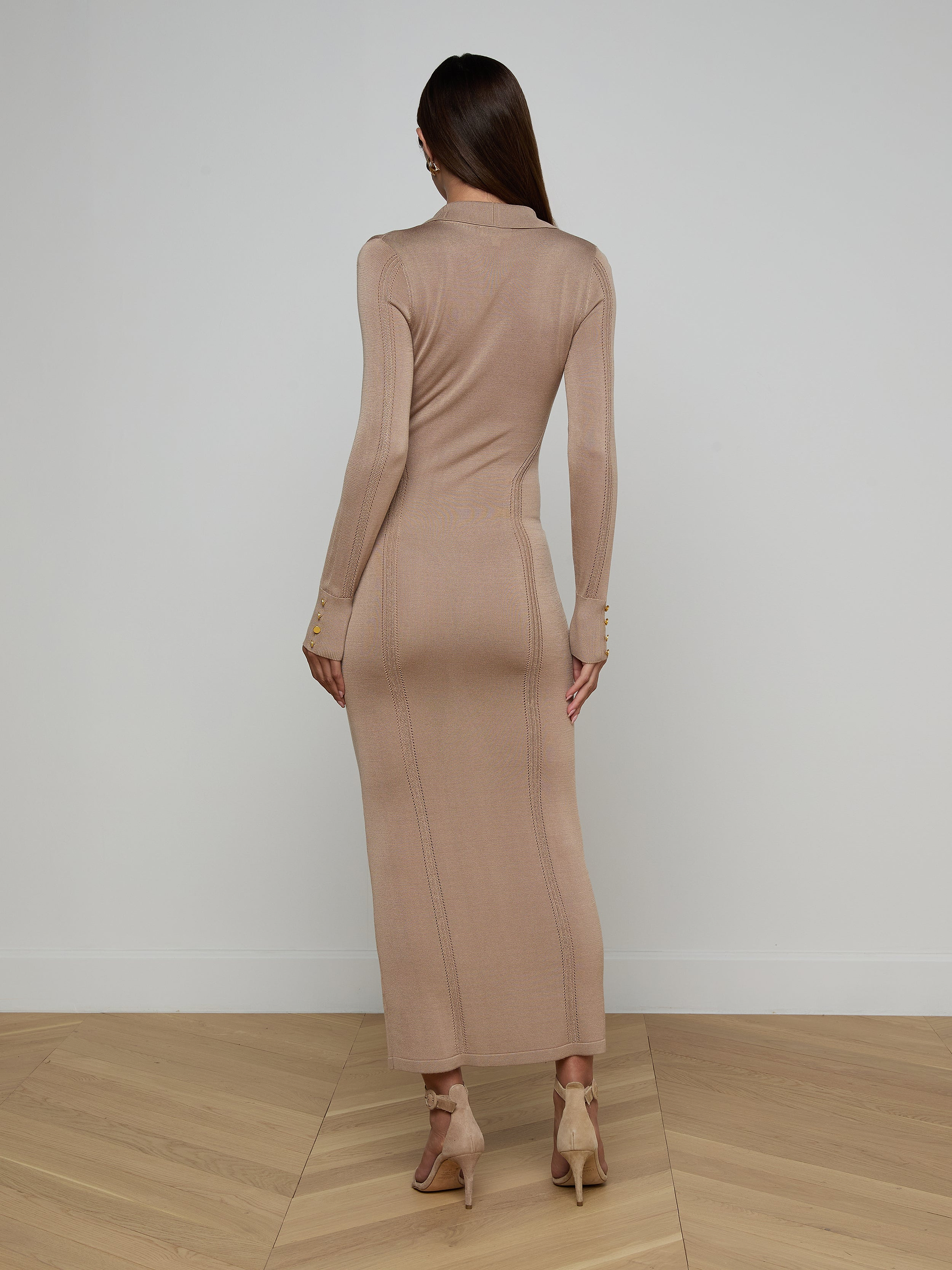 A woman stands facing away against a plain white background, her straight brown hair flowing down her back as she models the Delma Knit Maxi Dress—a long-sleeved, fitted knit blend—paired with beige heels.