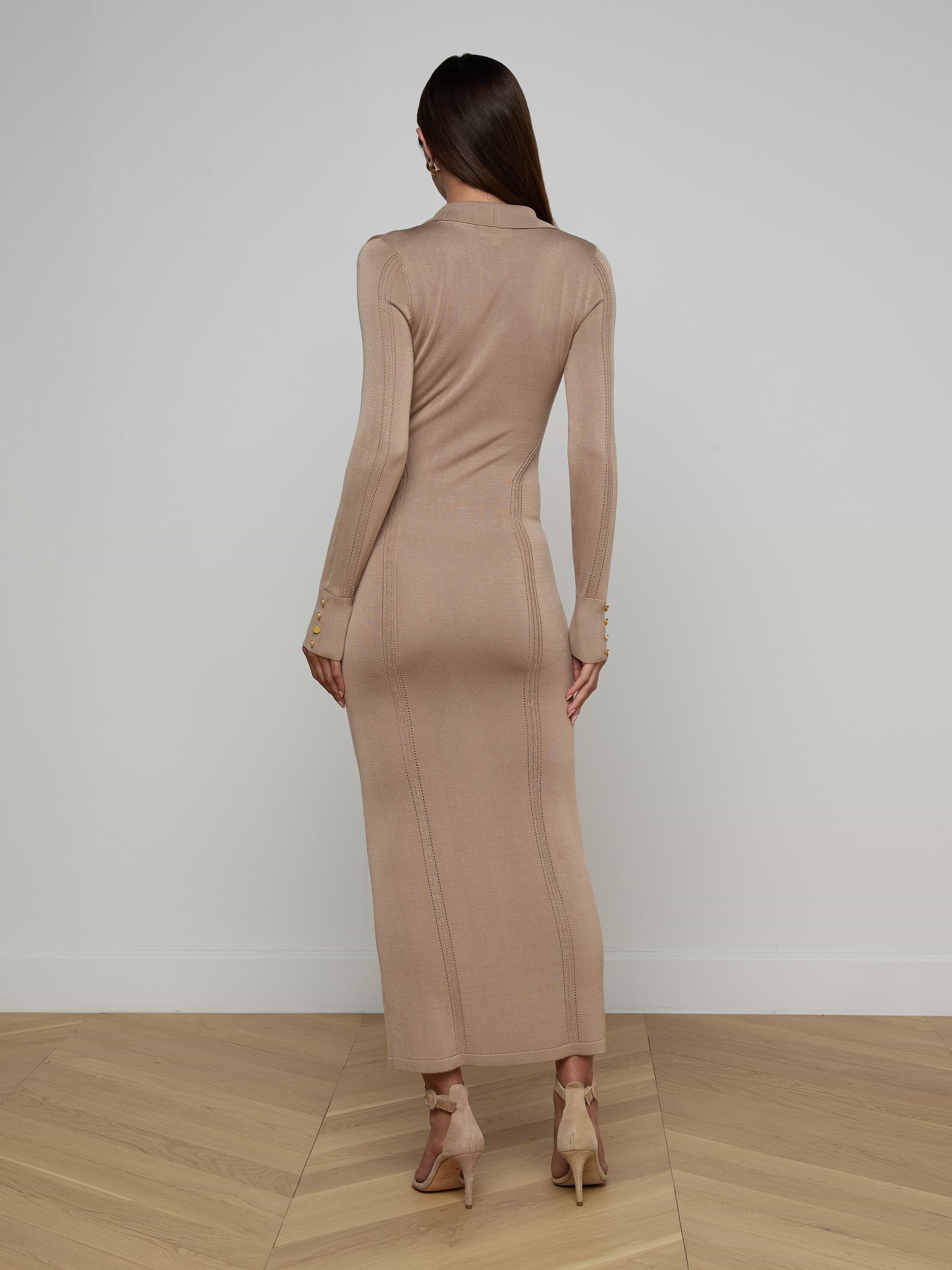 A woman stands facing away against a plain white background, her straight brown hair flowing down her back as she models the Delma Knit Maxi Dress—a long-sleeved, fitted knit blend—paired with beige heels.
