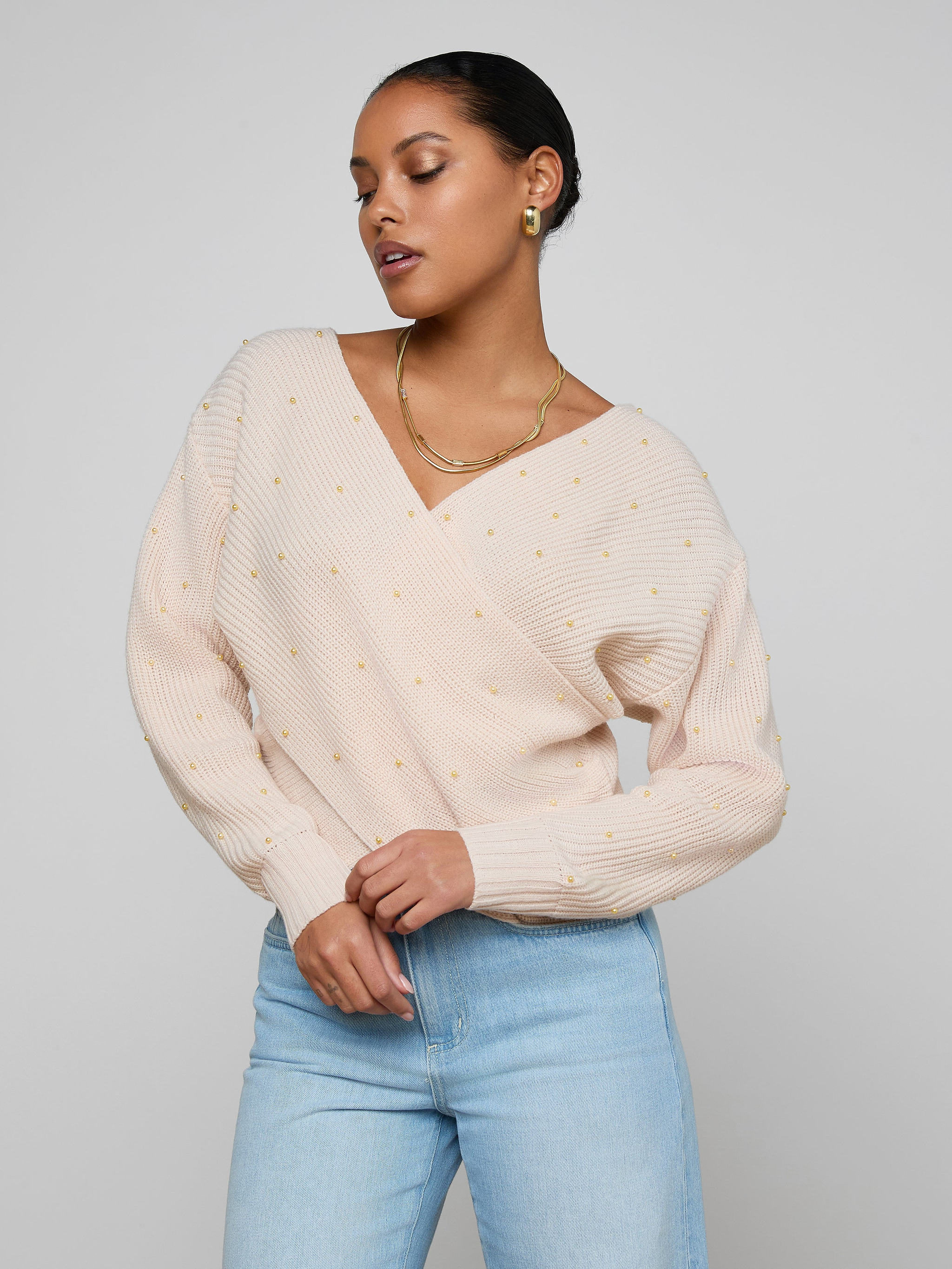 A woman wearing the Antonella Crossover Sweater—a light-colored, oversized wool knit with subtle dot patterns—paired with light blue jeans and gold jewelry, stands against a plain background in a relaxed pose with her head turned to the side.